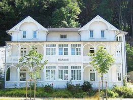 Weißes Gebäude im Bäderstil mit Veranda, umgeben von Bäumen.