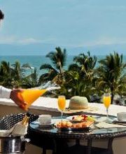 Hombre sirviendo jugo en mesa con desayuno frente al mar y palmeras.