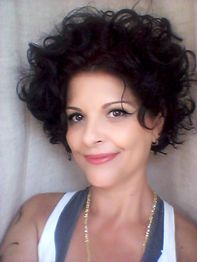 Mulher com cabelo cacheado escuro e blusa branca sorri para a câmera em um fundo neutro.