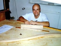 Ein Mann arbeitet an einem Holztisch mit Holzstücken und Plänen in einem Raum.