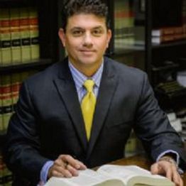 Man in a suit with a yellow tie reading a book, sitting at a table with shelves of books behind him.