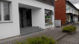 Eingang eines modernen Gebäudes mit Glastür, daneben Tisch und Stuhl auf grauem Pflasterweg.