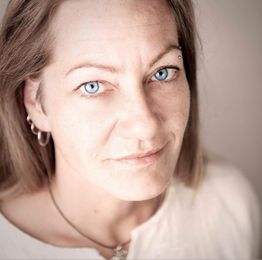 Porträt einer Frau mit langen Haaren, hellen Augen und Ohrringen, die in die Kamera schaut.