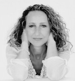 Eine Frau mit lockigem Haar hält ihr Gesicht in den Händen und schaut direkt in die Kamera.