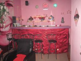 Kleine Bar mit roten, funkelnden Vorhängen, dekorierten Regalen und Barhockern vor pinker Wand.