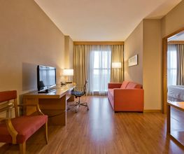 Sala de estar de hotel com sofá, mesa de trabalho, TV e vista para quarto com cama.