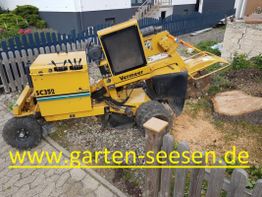 Gelbe Baumstumpffräse im Garten, umgeben von Holzzaun und städtischer Umgebung.