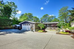 Single-story gray house with a wide driveway, surrounded by lush trees and a well-kept garden.