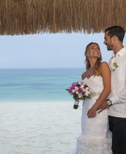 Pareja de boda sonríe en la playa bajo un techo de paja, con el mar de fondo.