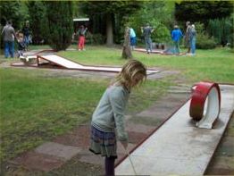 Meisje speelt minigolf op grasveld terwijl mensen op de achtergrond toekijken.
