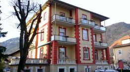 Dreistöckiges Hotel mit Balkonen, gelb-rot, in bergiger Umgebung, Baum im Vordergrund.