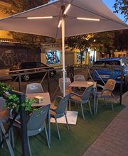 Terraza con mesas y sillas grises bajo una sombrilla blanca en una calle con coches estacionados.