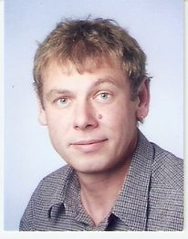 Ein Mann mit hellem Haar und kariertem Hemd lächelt leicht in die Kamera.