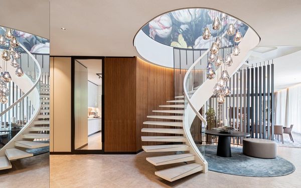 Wendeltreppe aus Stahl hell lackiert mit einem Glasgeländer mündet in einer kreisförmigen Öffnung ins nächste Obergeschoss. Es hängen Pendelleuchten zur Deko in der Mitte der Öffnung.