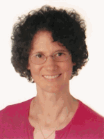 Person mit lockigem Haar und Brille, lächelt, trägt ein rosa Oberteil.