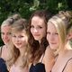 Vier junge Frauen in eleganten Kleidern draußen, im Hintergrund grüne Blätter.