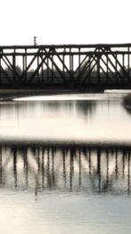 Eisenbahnbrücke spiegelt sich im ruhigen Flusswasser unter bewölktem Himmel.