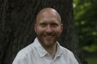 Ein lächelnder Mann vor einem Baum in einer grünen Umgebung trägt ein weißes Hemd.