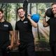 Drie mannen met sportkleding lachen in een fitnessruimte met een boksbal en gewichten.