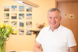 Ein lächelnder Mann in einem weißen Hemd steht neben gelben Tulpen und Bilderrahmen an der Wand.