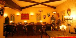 Ritterlicher Saal mit langen Tischen, Kerzenlicht, Rüstungen und an den Wänden Wappen.