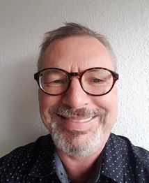 Person mit Brille lächelt, trägt ein dunkles Hemd. Hintergrund ist weiße, strukturierte Wand.