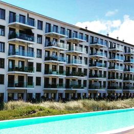Mehrstöckiges Wohngebäude mit Balkonen, davor ein Swimmingpool und Gräser.