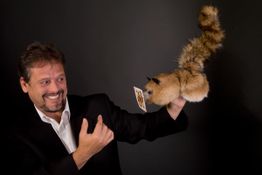 Mann lächelt Eichhörnchen-Puppe an, die eine Spielkarte hält, vor schwarzem Hintergrund.