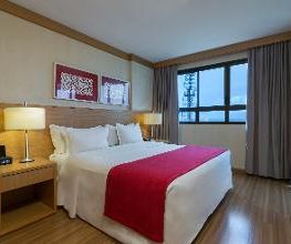 Quarto de hotel com cama de casal, roupa de cama branca, manta vermelha e janela com cortinas.