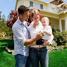 A smiling family holds a baby in front of a suburban house with a neat lawn and garden.