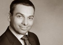 Mann in Anzug lächelt; er hat kurzes Haar und einen dezenten Hintergrund in Sepiaton.