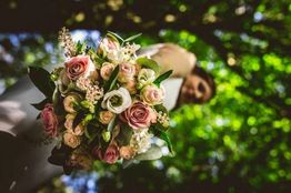 Bruid met boeket van roze en witte bloemen, kijkend door groene bladeren in buitenomgeving.