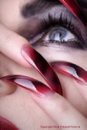 Nahaufnahme eines Auges mit betonten Wimpern und langen, rot glänzenden Fingernägeln.