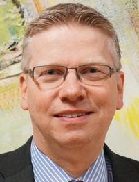 Mann mit Brille und Anzug lächelt vor einem farbenfrohen Hintergrund.