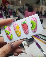 Uñas postizas decoradas con frutas: aguacate, limón, fresa y kiwi, sobre fondo rosa.