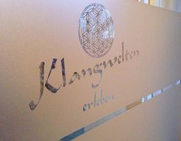 Logo mit der Aufschrift "Klangwelten erleben" auf einer Wand; darüber geometrisches Muster.