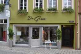 Grünes Gebäude mit Ladenfront, großen Fenstern und Pflanzen auf dem Gehweg.