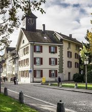 Straßenszene mit historischem Gebäude und Uhrenturm bei Herbstwetter. Personen spazieren entlang.