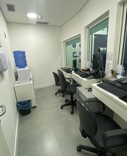 Sala de escritório com mesas, cadeiras, computadores e bebedouro ao fundo.