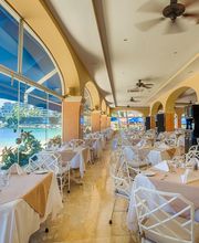 Restaurante con mesas elegantes junto a grandes ventanas con vista al mar y decoración tropical.