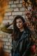 Woman in a leather jacket poses by a brick wall with autumn leaves.