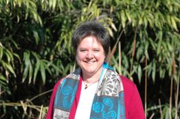 Eine lächelnde Frau steht vor grünem Blattwerk, trägt einen Schal und eine rote Jacke.