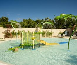 Piscina infantil con juegos de agua y tobogán bajo un cielo azul claro.