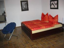 Schlafzimmer mit rotem Bett, blauer Stuhl, Fliesenboden und zwei Wandbildern.