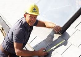 Smiling worker in a yellow hard hat measures a rooftop with a tape measure.