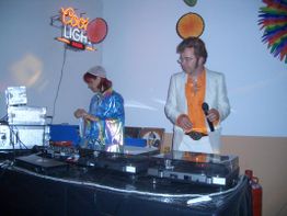Zwei DJs stehen an einem Mischpult, umgeben von bunter Partydeko und einem "Cool Light" Schild.
