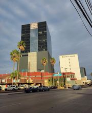 Edificio alto con letrero de venta/renta al lado de un hotel; palmeras y autos en frente.