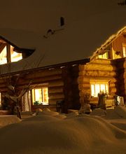 Verschneites Blockhaus bei Nacht, warme Beleuchtung im Inneren, dicker Schnee bedeckt das Dach.