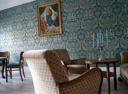 Wohnzimmer mit gemusterter Tapete, Gemälde, Polstersesseln, Tisch und Kerzenständer.