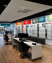 Optiekwinkel met brillen aan de muur, bureaus en stoelen op een houten vloer.